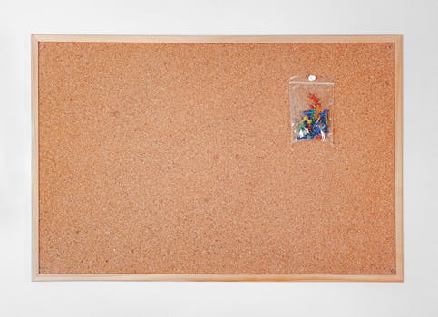 Cork board with wooden frame featuring a packet of colorful push pins for office or home use.