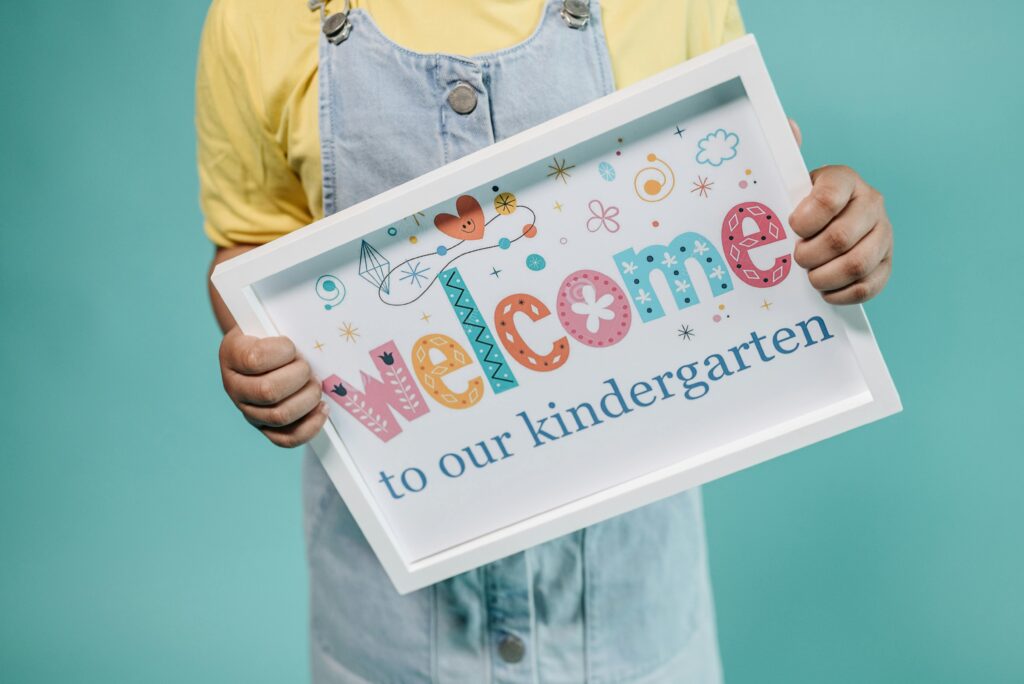 A child holding a colorful welcome to kindergarten poster against a teal background.